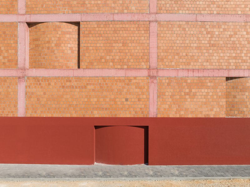 Curved red entry door set into red plinth below terracotta brick facade with arched openings