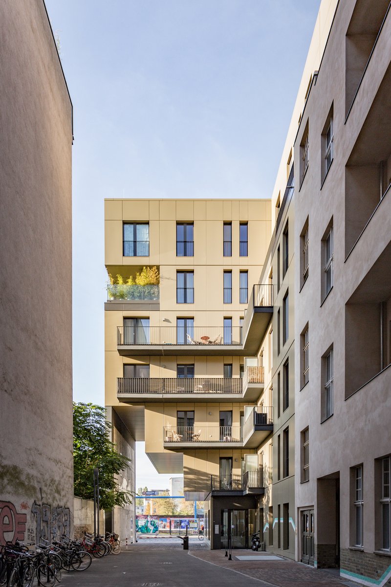 Street view framed by adjacent buildings showing the golden-paneled facade with protruding balconies and bicycles below