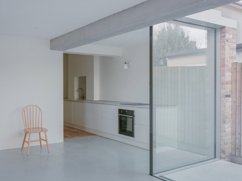 Minimalist kitchen with grey cabinetry and single wooden chair beside floor-to-ceiling glass overlooking timber screening