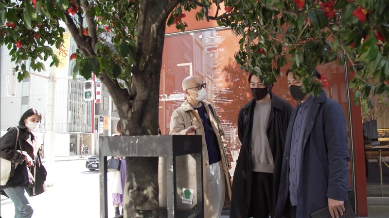 Four people gathered beneath a tree outside a glass-fronted exhibition space in daylight