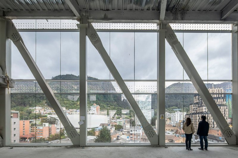 Full-height window wall with diagonal white structural bracing as two people overlook the surrounding cityscape