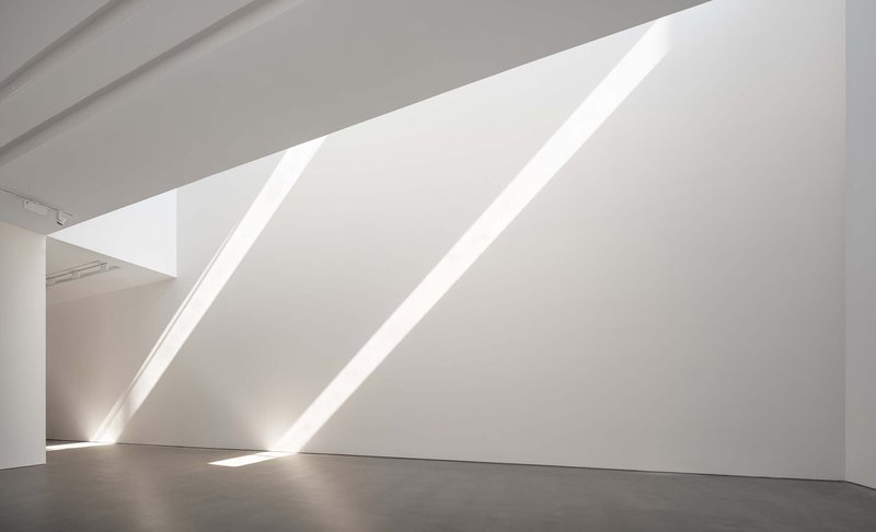 White gallery wall with diagonal skylight slots casting sharp shadows across the polished floor