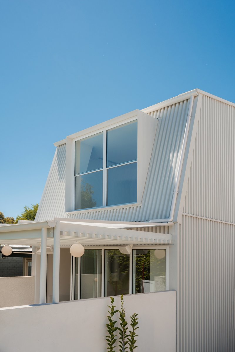Two-story white corrugated facade with angled upper window and covered entry pergola below clear blue sky