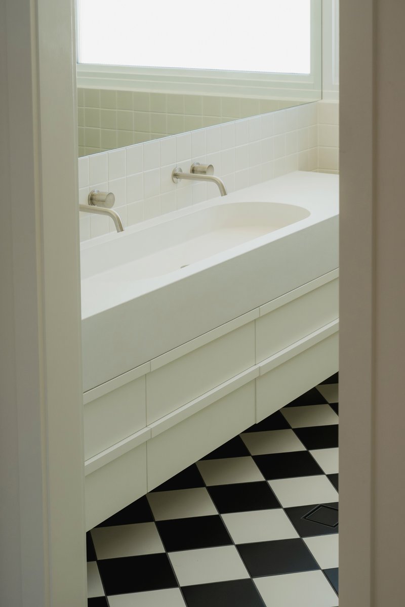 View through doorway to vanity with wall-mounted taps and checkerboard floor in natural daylight