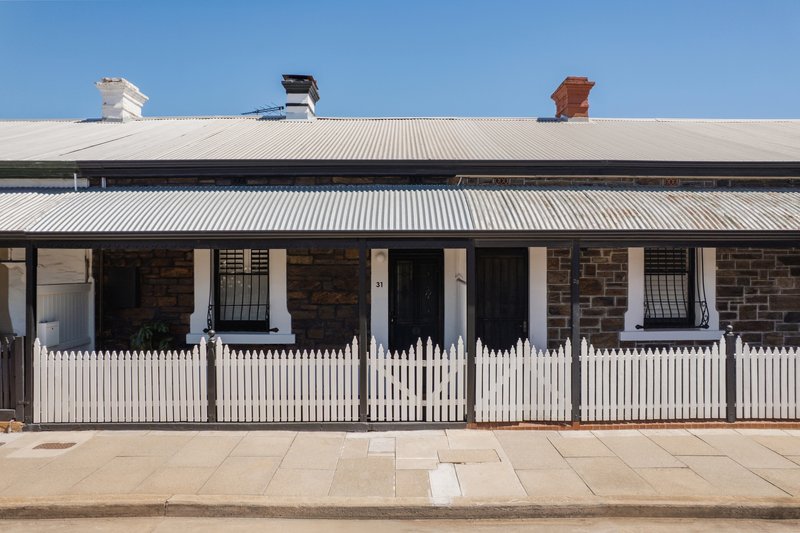 Street view of a stone cottage with corrugated metal roof, white picket fence and verandah under clear blue sky