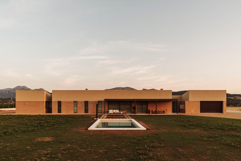 Rear elevation with rammed earth walls and rectilinear swimming pool surrounded by open lawn at dusk