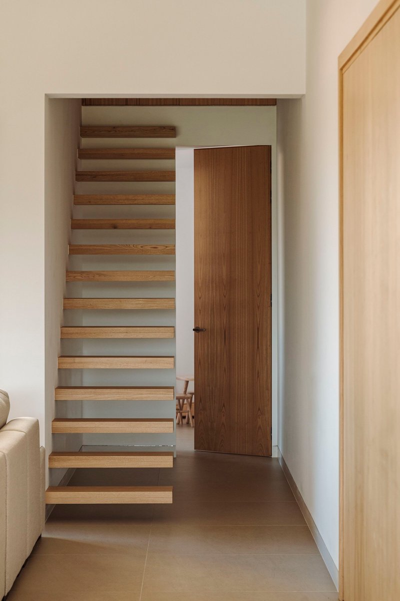 Floating timber staircase with open risers rising beside a closed wooden door in a white hallway