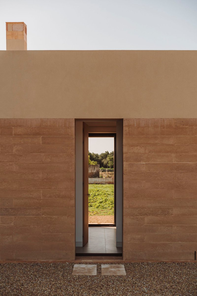Tall narrow doorway framing view of gravel courtyard and green lawn beyond rammed earth facade