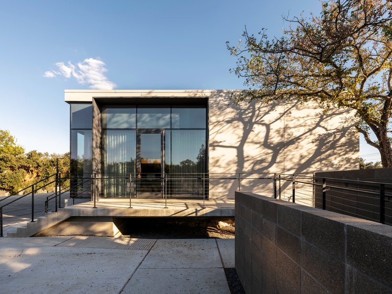 Glass-enclosed entrance with metal railing overlooking a stone-clad retaining wall and tree shadow