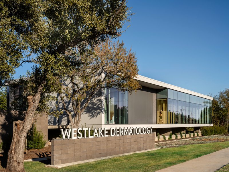Entry view with stone base wall and glass facade set behind established oak trees