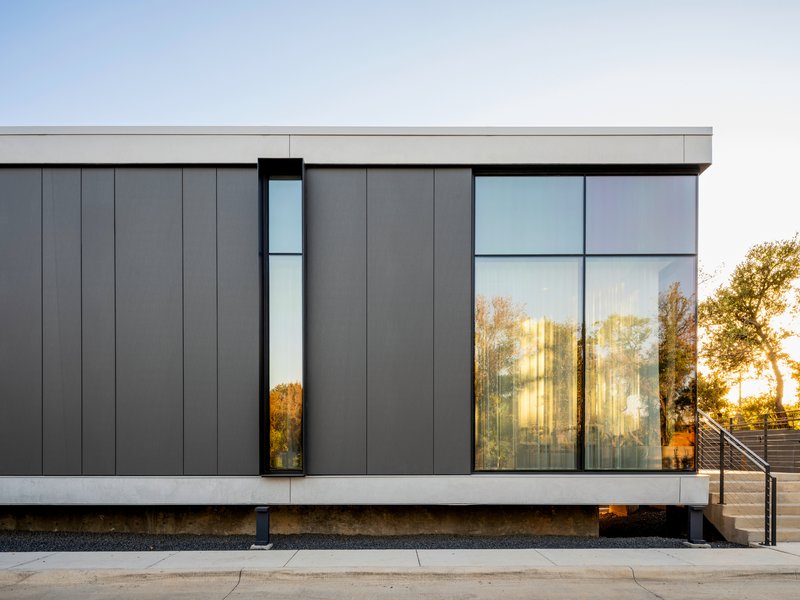 Dark metal panel facade with narrow vertical window and full-height glazing reflecting surrounding trees at dusk