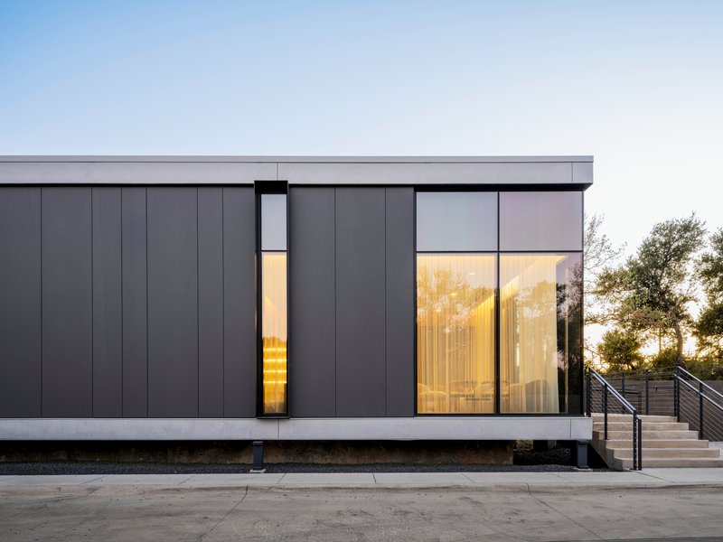Dark paneled facade with narrow vertical slot window and illuminated interior visible through floor-to-ceiling glass