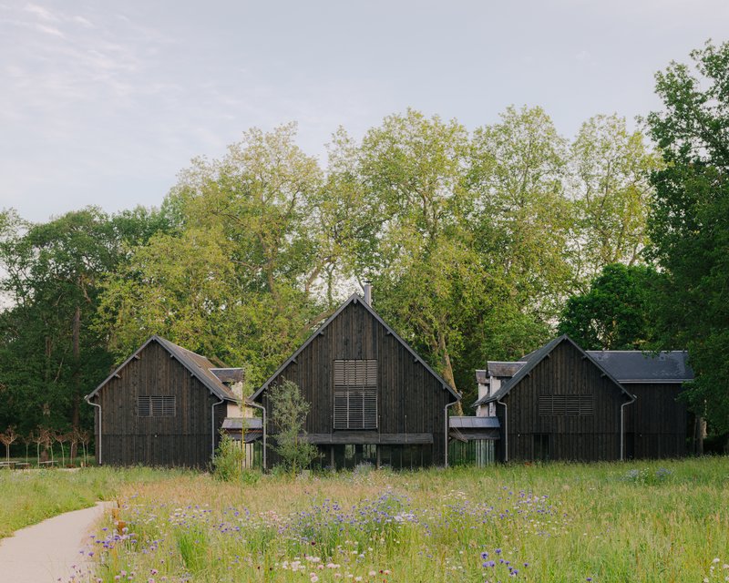 Three gabled timber structures with shuttered openings set in a wildflower field with trees behind