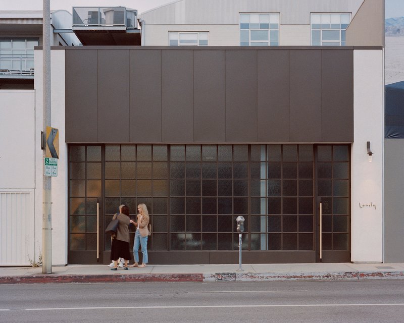 Street facade with gridded glass storefront and dark metal panel upper volume as two pedestrians pause at the entrance