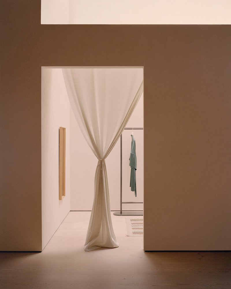Doorway framed in warm brown walls with draped linen curtain tied back, revealing clothing rack beyond