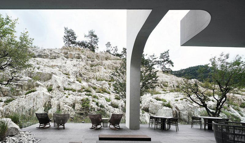 Covered terrace with seating and dining furniture facing the exposed rock outcrop beyond curved concrete columns