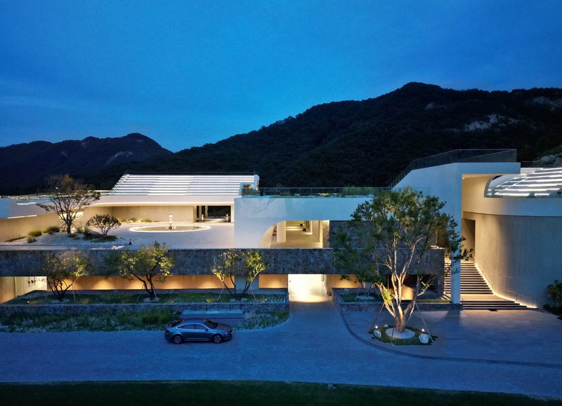 Night view of the white horizontal volumes with ribbed roofs and illuminated openings beneath the dark mountains