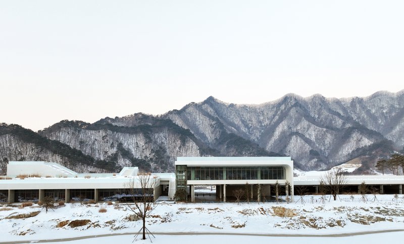 Low horizontal volumes with flat roofs set in a snowy field with bare trees and mountains behind