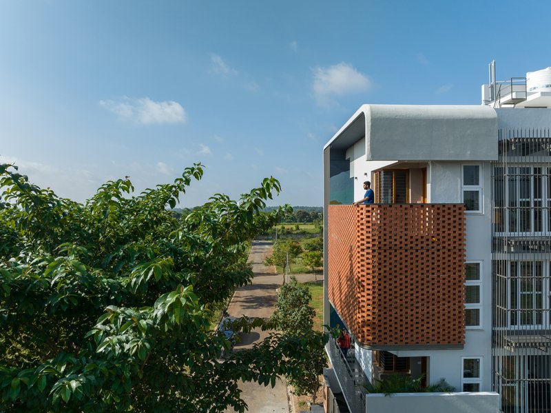 Perforated terracotta screen wrapping a projecting balcony where a figure stands overlooking the green surroundings