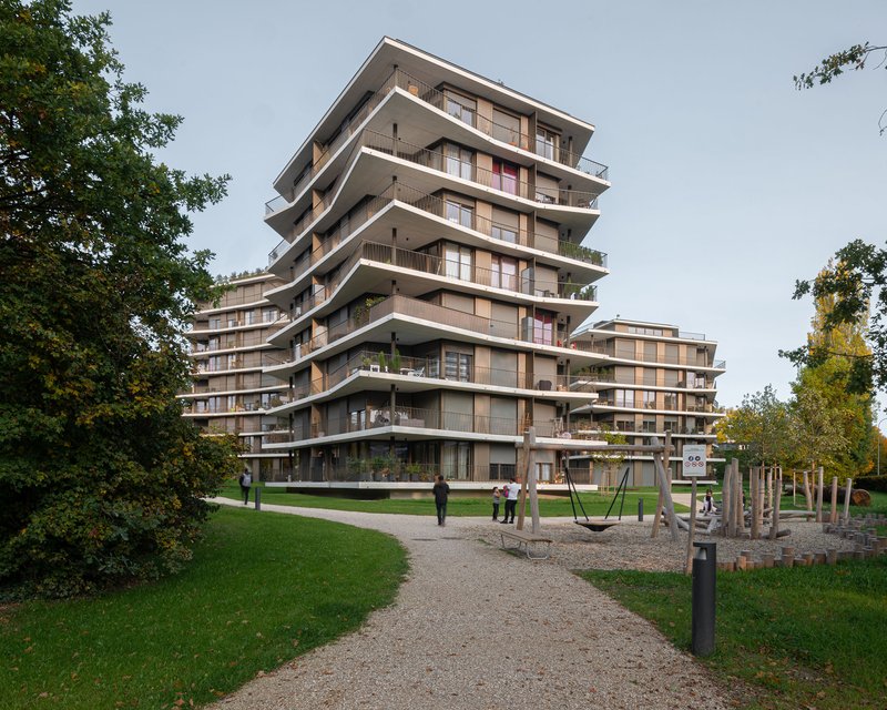 Stepped residential tower with deep concrete balconies and timber screens seen from a park pathway