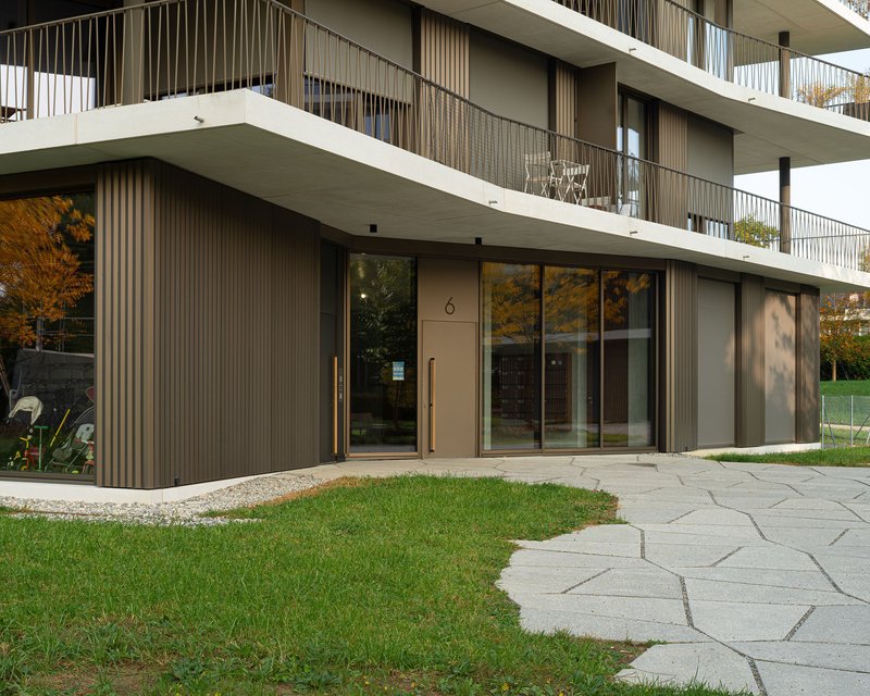 Ground level facade with ribbed metal panels, glass entry door, and irregularly jointed stone paving in sunlight
