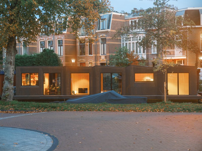 Street view of the houseboat's illuminated glazed facade beneath the brick residential buildings at twilight