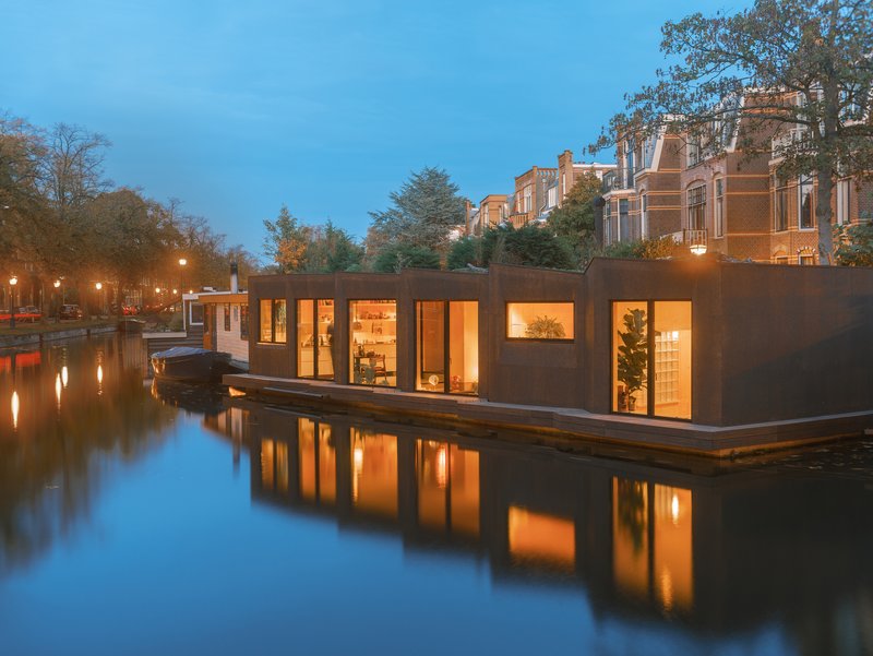 Linear facade of the floating dwelling with large glazed openings along the timber deck at twilight