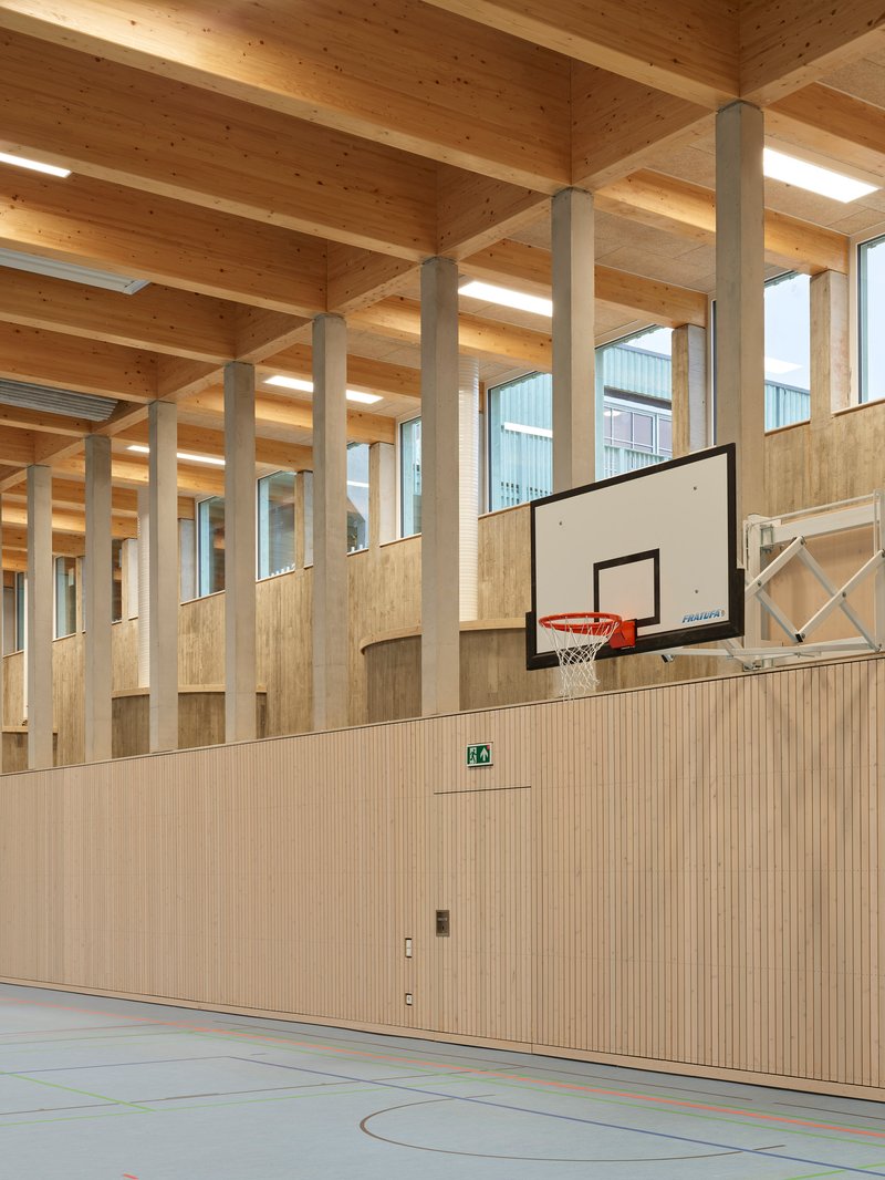Gymnasium with basketball hoop against slatted wood storage wall beneath exposed timber beam ceiling and columns