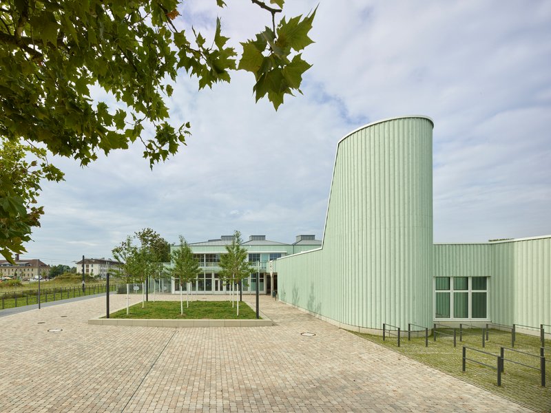 Paved courtyard leading to cylindrical corrugated metal tower with young trees along pathway under overcast sky