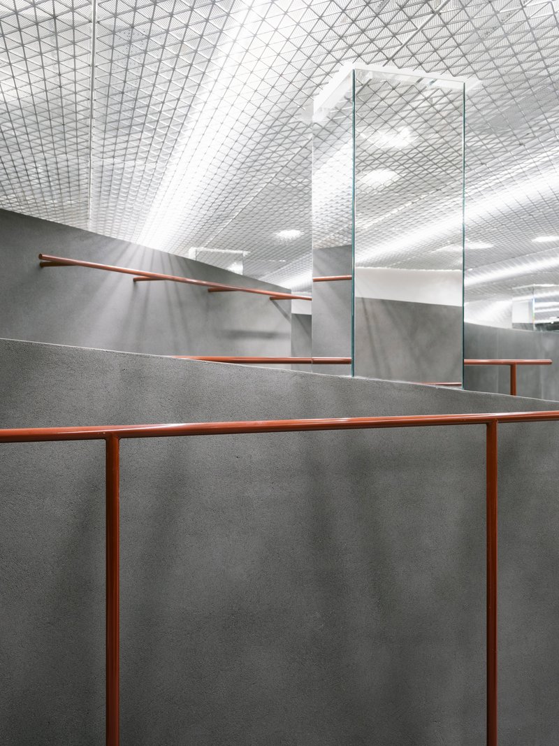 Interior view of grey concrete partition walls with copper handrails beneath a diamond-patterned mesh ceiling