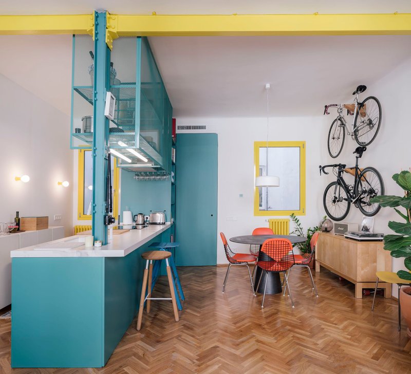 Open living area with teal kitchen island, yellow structural beam, and wall-mounted bicycles above herringbone flooring