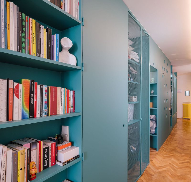 Teal shelving unit with books and perforated metal cabinet doors along a herringbone parquet corridor