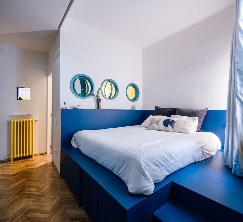 Bedroom with blue platform bed, three circular porthole windows, and yellow radiator against white walls