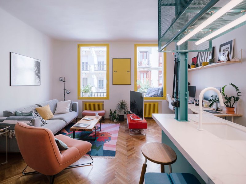 Open living area with yellow-framed windows, teal kitchen island and herringbone wood flooring in daylight