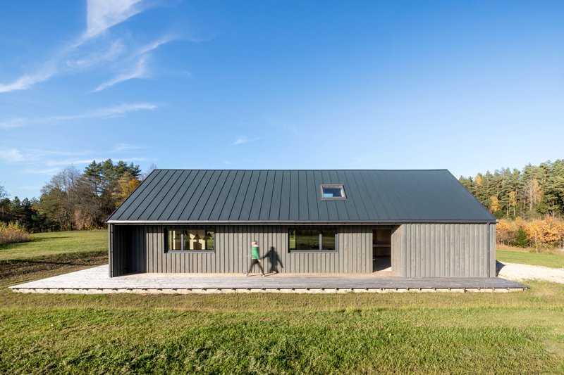 Side elevation with vertical timber cladding and standing-seam metal roof set in an open meadow under clear blue sky