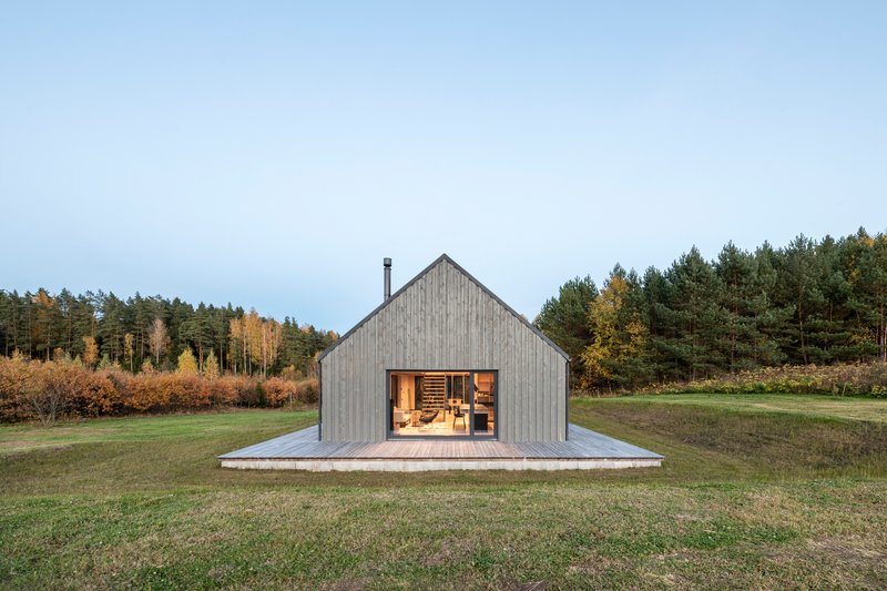 Gabled end facade with full-height glazing and timber deck surrounded by meadow and forest
