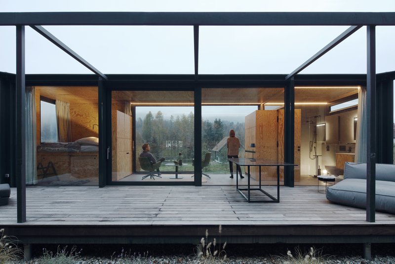 Timber deck terrace beneath steel pergola framing views into plywood-clad rooms with two people in misty weather