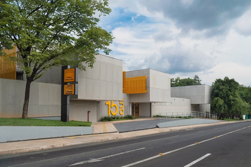 Street facade showing concrete volumes, yellow metal box, and mature trees framing the entrance