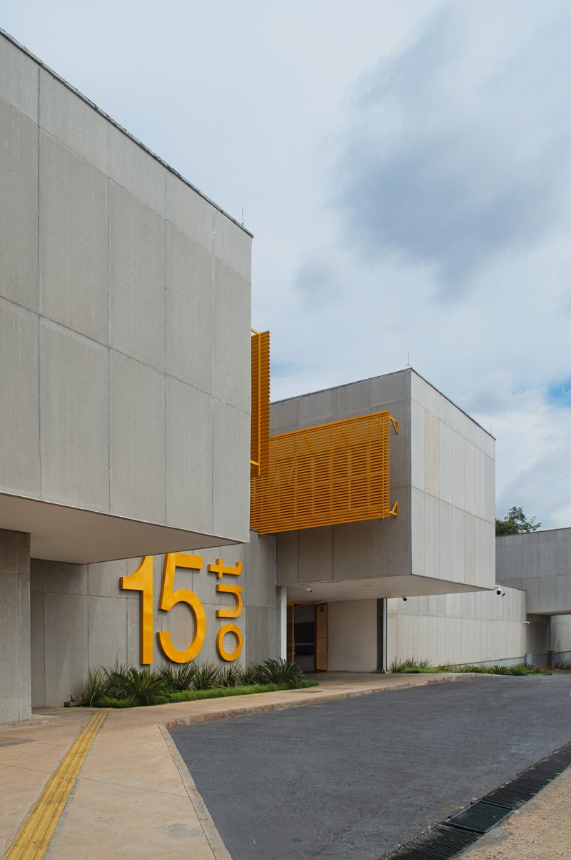 Street entrance with concrete facade, yellow metal louvers, and large dimensional numerals beside planted beds under cloudy sky
