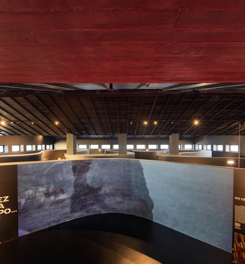 Interior gallery space with curved projection screen beneath an exposed slatted ceiling and crimson vault above
