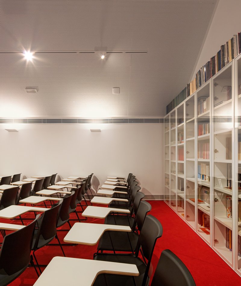 Classroom with rows of tablet-arm chairs on red carpet and a built-in bookshelf under perforated ceiling panels