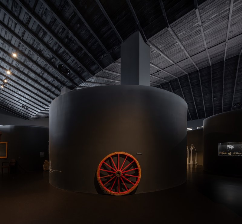 Cylindrical museum display volume with a red wagon wheel at its base beneath an exposed timber ceiling