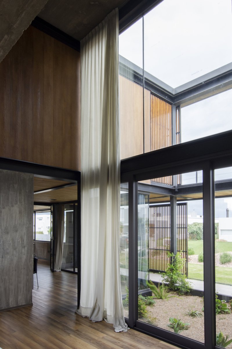 Double-height interior corner with floor-to-ceiling glazing, timber screens, and a planted courtyard visible beyond