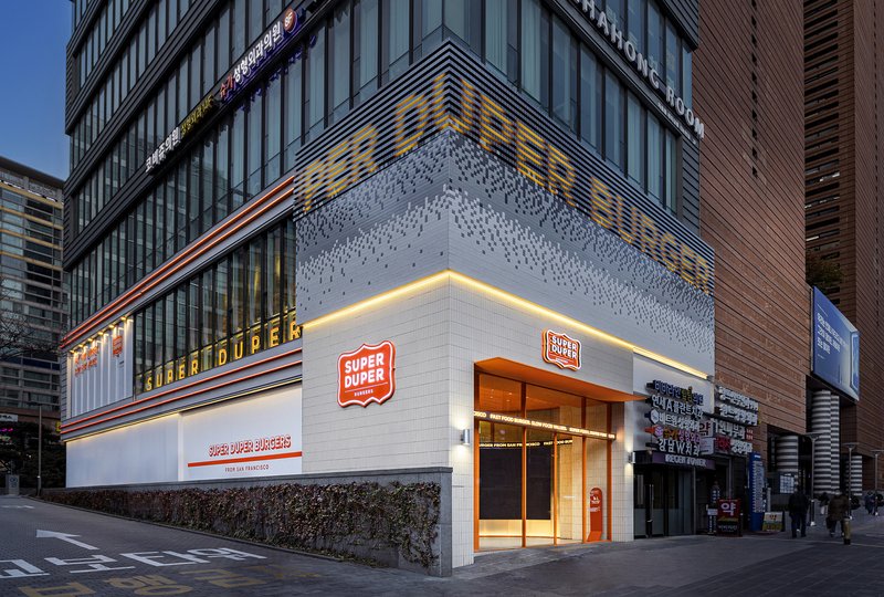 Street corner facade with white tiled lower volume and textured metal upper section at dusk