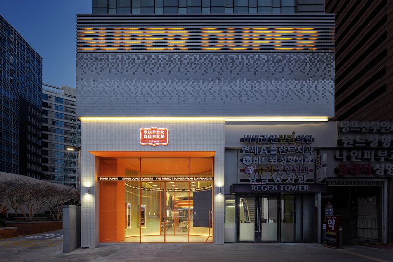 Entrance facade with orange metal doors and illuminated signage at twilight between neighboring buildings