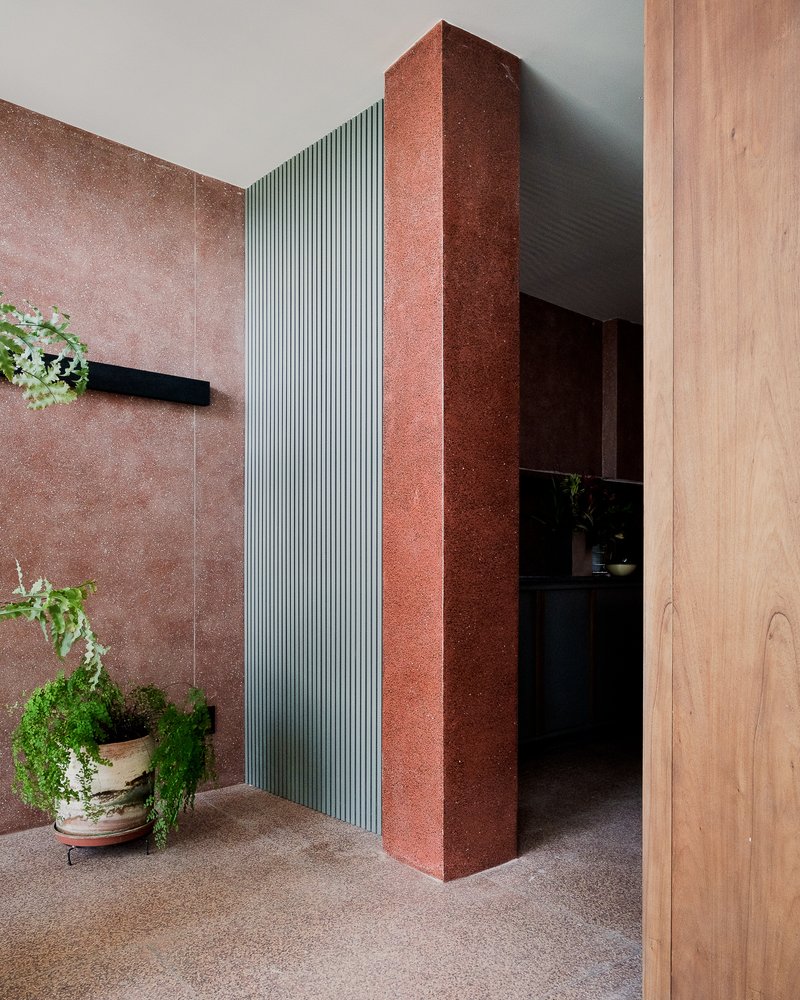 Entrance corridor with red textured stone column, ribbed metal panel and potted ferns on terrazzo floor