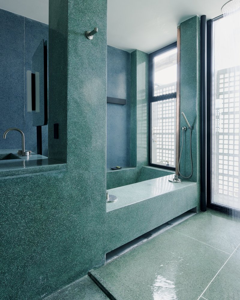 Bathroom with teal terrazzo walls, recessed bathtub and black-framed window opening to a glass brick screen
