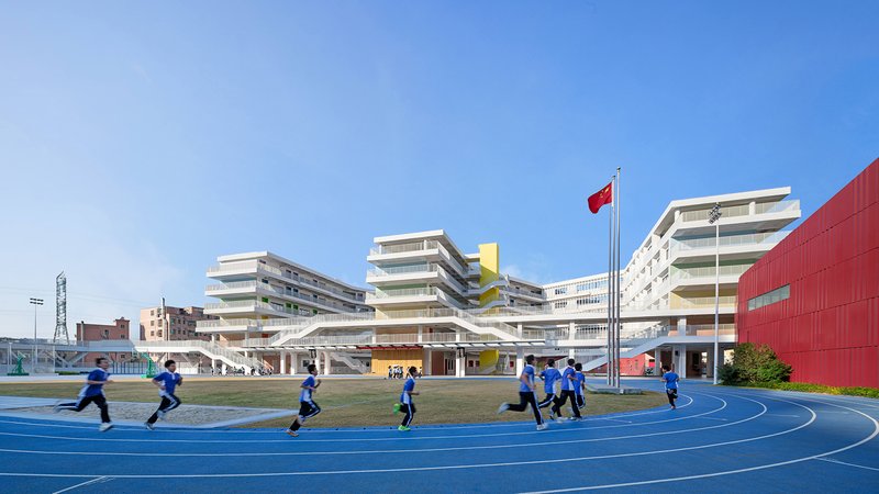 Layered white school buildings with horizontal balconies overlooking a blue running track with students in motion