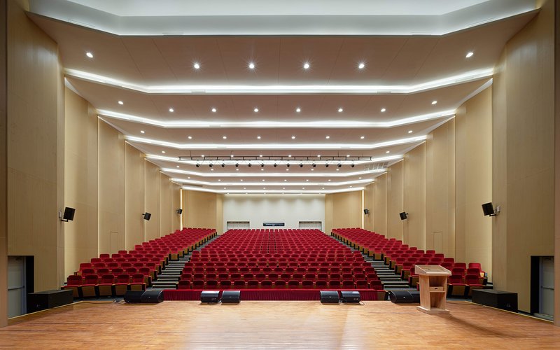 Auditorium interior with tiered red seating, wooden stage, and recessed lighting panels in curved ceiling coffers