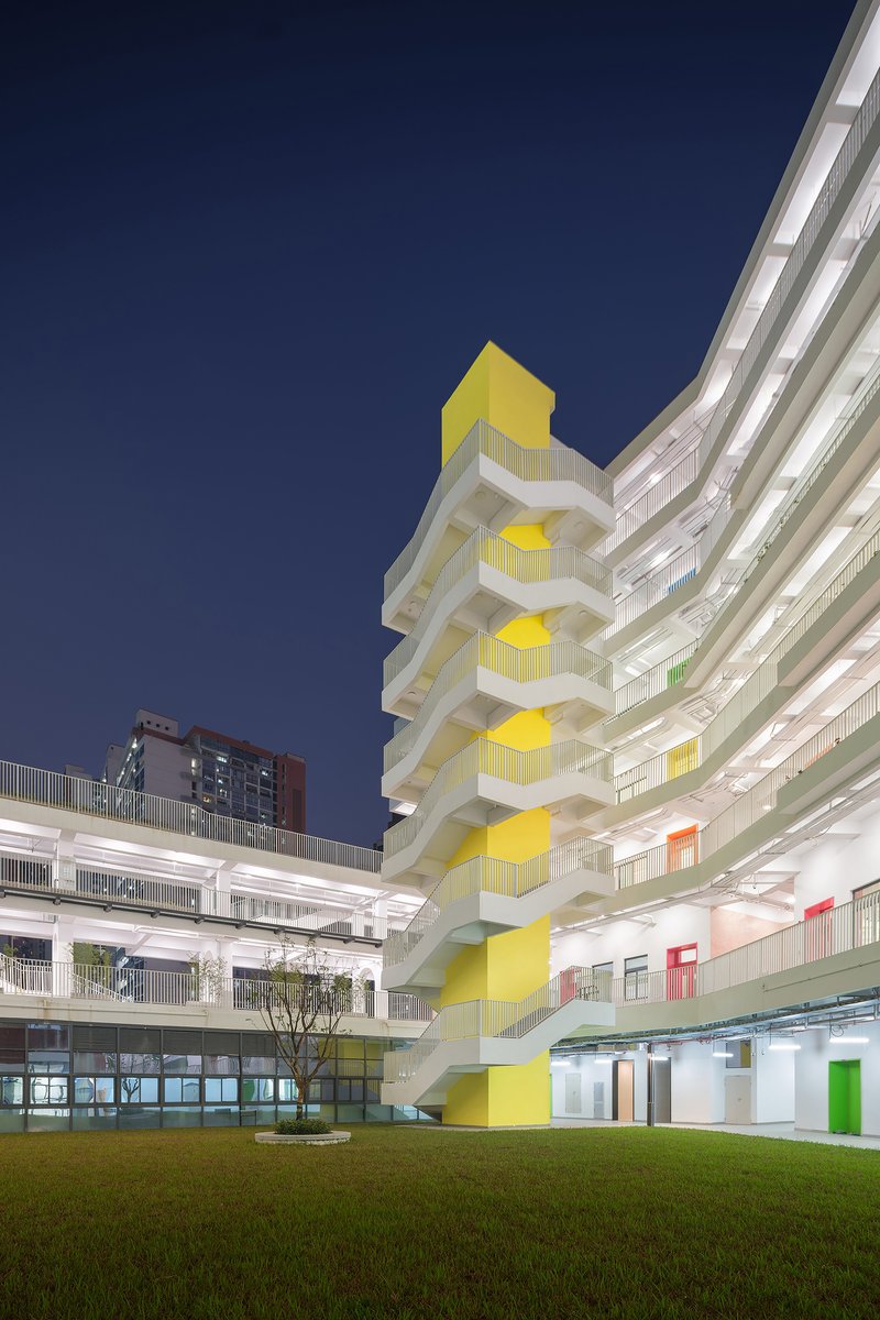 Yellow stacked staircase tower illuminated against a dark blue evening sky beside white terraced school buildings